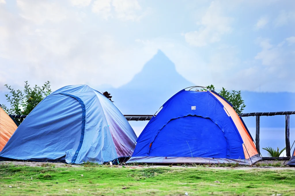Two camping tents, one blue and orange, and another blue, set up near a lake with a mountain in the background.