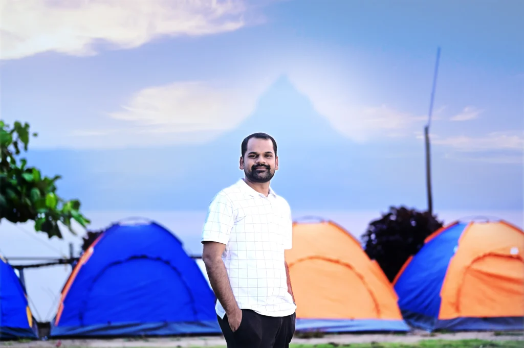 A man in a white checkered shirt standing in front of blue and orange camping tents.