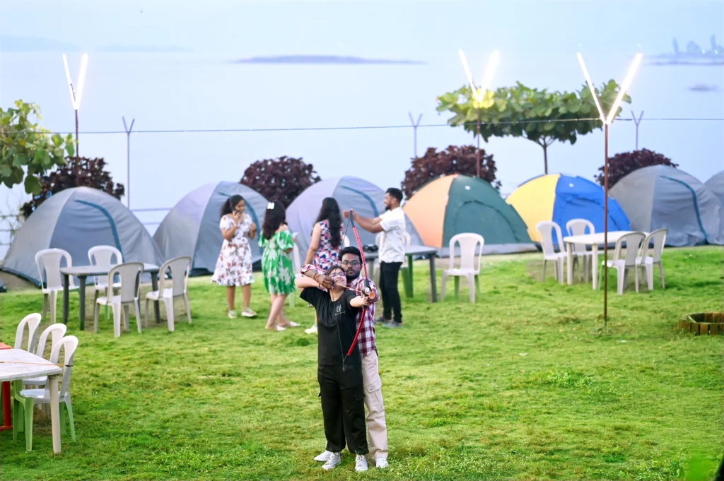 Two individuals posing for a photo at a camping site with tents and people in the background.